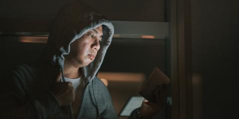 Een man zit in een donkere kamer, heeft zijn capuchon van zijn hoodie op en kijkt naar een tablet. Zijn gezicht is zichtbaar door het licht van de tablet.