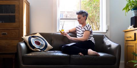 Vrouw zit thuis op haar bank voor het raam. Ze heeft haar telefoon vast en kijkt bezorgd.
