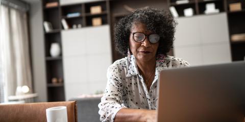 Een senior vrouw zit thuis achter haar laptop. Ze draagt een bril en heeft zwart haar.