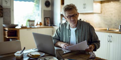 Senior man zit aan keukentafel met brief rekening in hand en laptop voor zich