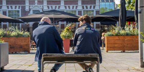 Twee medewerkers van de Nationale ombudsman zitten op een bankje in de zon (gefotografeerd in de rug). 