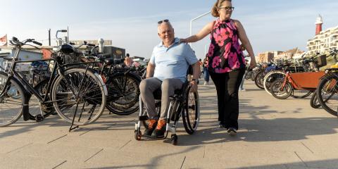 Ouder stel loopt langs het strand. Een vrouw heeft haar hand op de schouder van haar partner, die in een rolstoel zit