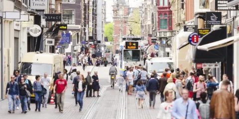 Een foto van een straat in Amsterdam. Je ziet mensen wandelen, tramrails, en aan beide kanten winkels.