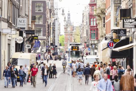 Foto van een straat in Amsterdam. Je ziet wandelende mensen, tramrails en winkels aan beide zijden.