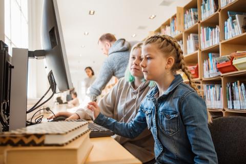 Twee jonge meisjes kijken naar het computerscherm. Ze zitten in een bibliotheek