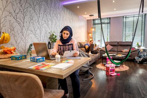 Vrouw zit aan tafel in de woonkamer. Ze kijkt in haar laptop. Op de grond ligt speelgoed