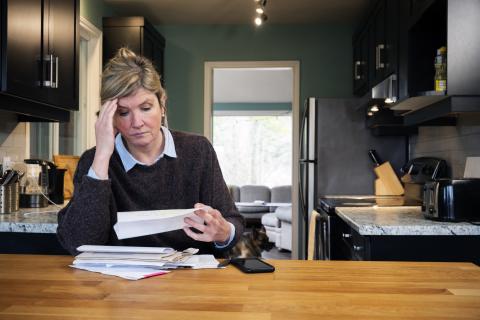 Een vrouw zit aan tafel en kijkt zorgelijk in papieren. Haar hand ondersteund het hoofd