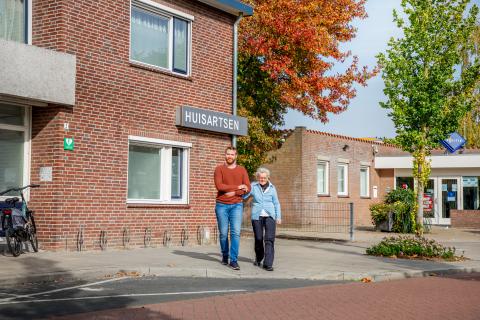 Oudere dame wordt ondersteund tijdens het lopen door een jongere man. Ze lopen op straat