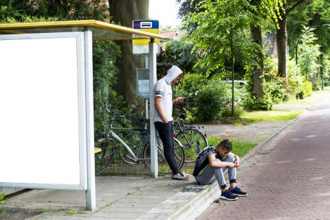 Twee jongens staan bij een bushokje. Ze kijken op hun telefoon