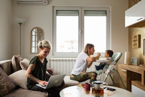 Een vrouw zit op de bank en werkt op haar laptop. Een andere vrouw geeft een kind te eten