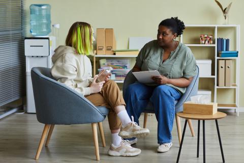 Twee vrouwen zijn in gesprek. Ze zitten in een blauw gekleurde stoel