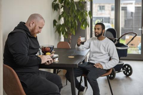 Twee mannen zitten aan een tafel. In de kamer staat een plant en kinderwagen