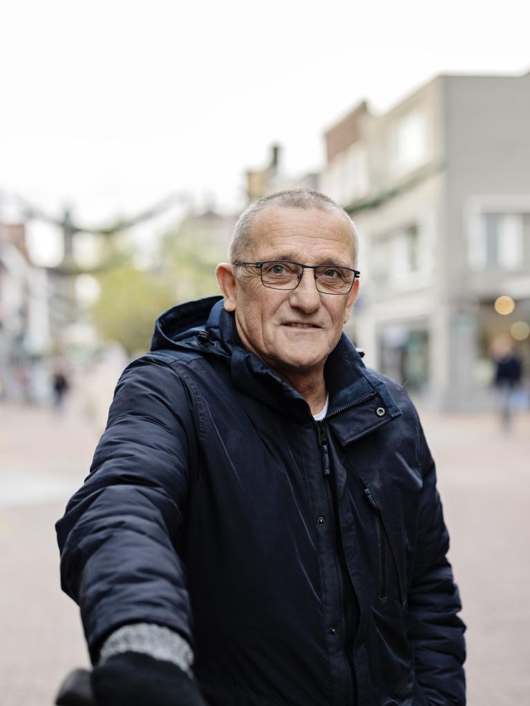 Senior man met bril en donkerblauwe jas staat op straat en kijkt in de camera.