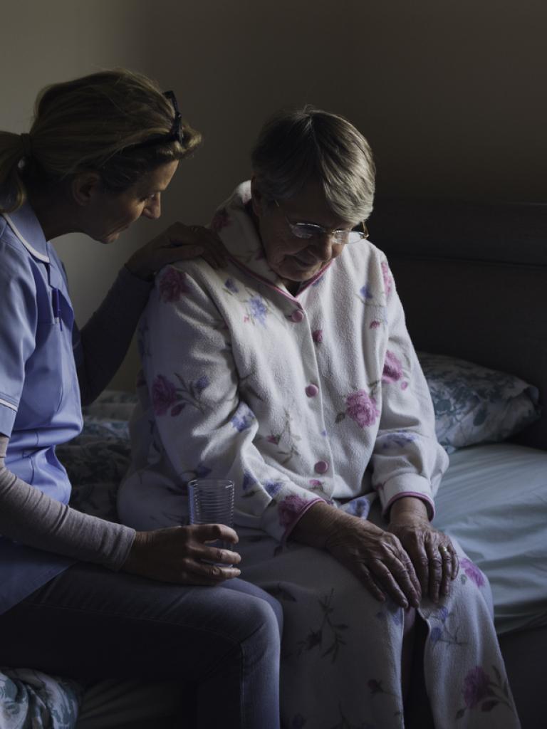 Oudere dame zit op bed zit samen met een verpleegster. De verpleegster slaat een arm om de vrouw heen.