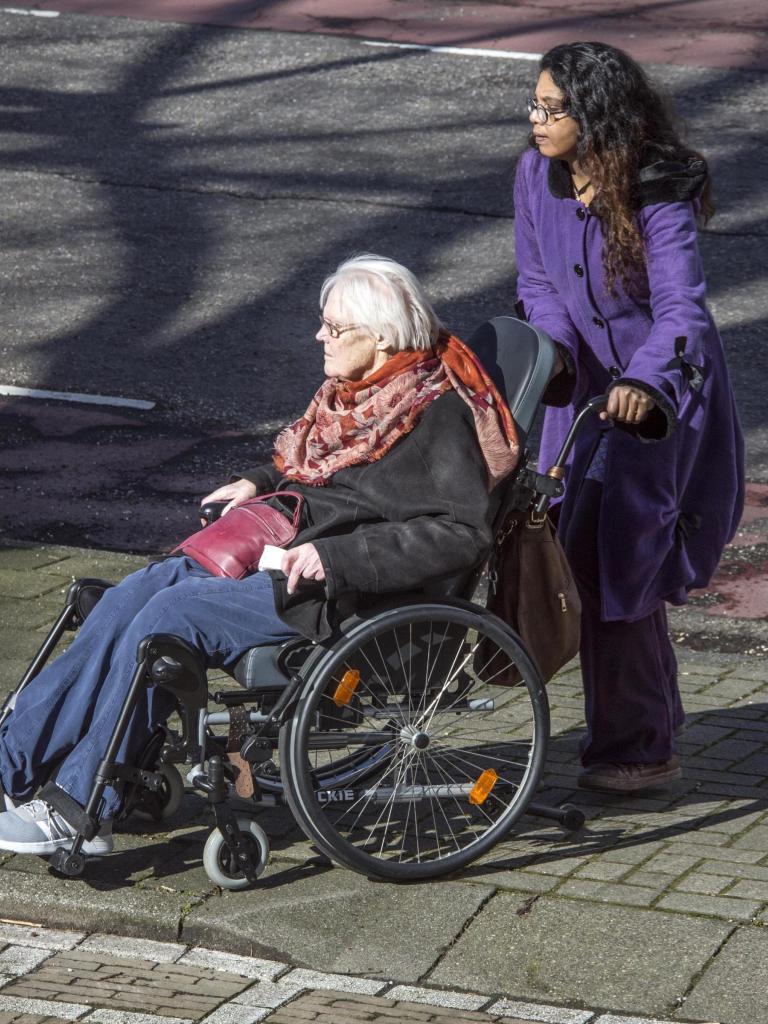 Mantelzorger (vrouw, lang donker haar en paarse jas) duwt rolstoel oudere vrouw (grijs haar, bril, donkere jas en rode sjaal
