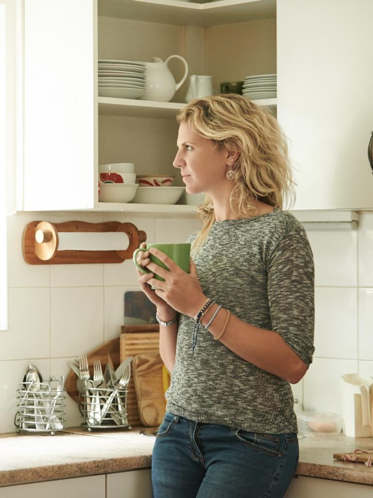 Een vrouw met blond krullend haar staat in haar keuken en kijkt uit het raam. In haar hand heeft ze een mok.