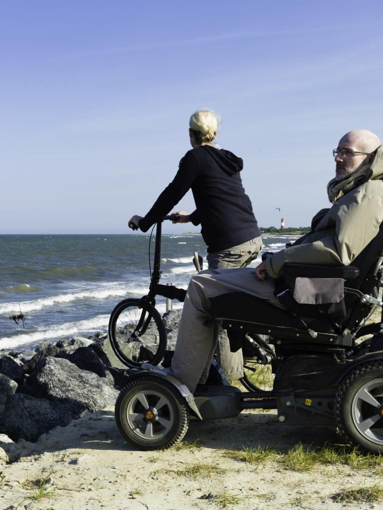 Een senior man zit in een scootmobiel. Naast hem staat een vrouw met een fiets. Ze staan op de rotsen dicht bij zee.
