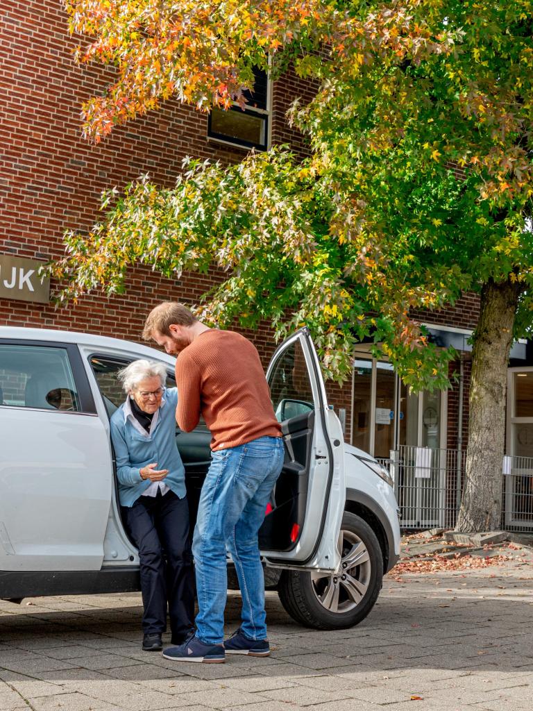 Mantelzorger helpt oudere vrouw uit de auto. Ze staan geparkeerd voor de huisartsenpraktijk.