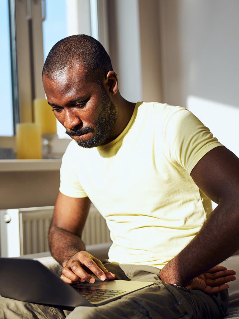 Een jongvolwassen man met geel t-shirt zit op zijn bank en heeft een laptop op zijn schoot. Hij kijkt bedenkelijk.