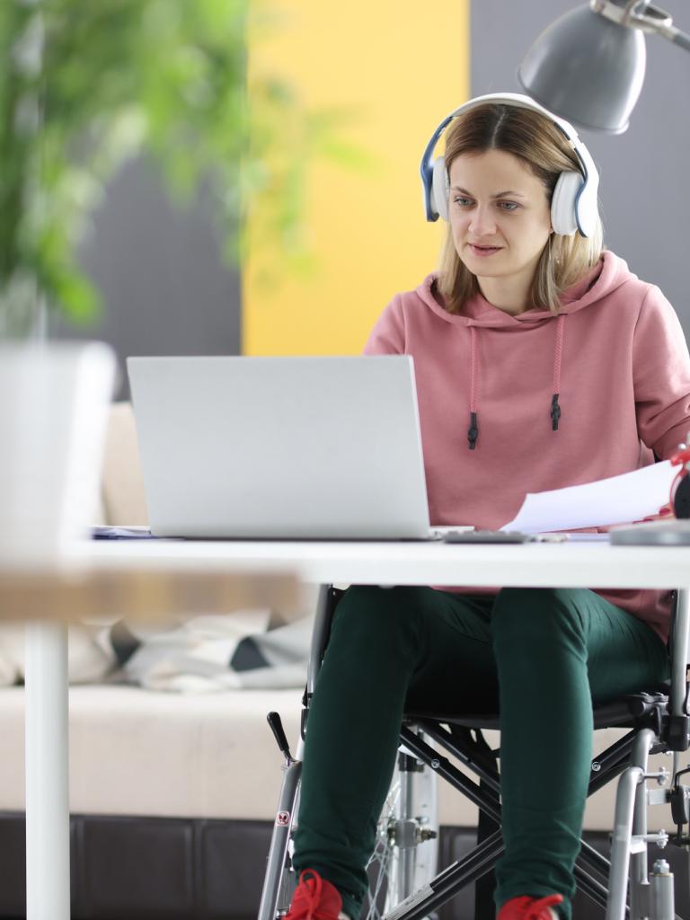Een jongvolwassen vrouw in een rolstoel zit thuis achter haar laptop te werken of studeren. Ze draagt een roze trui en heeft een koptelefoon op.