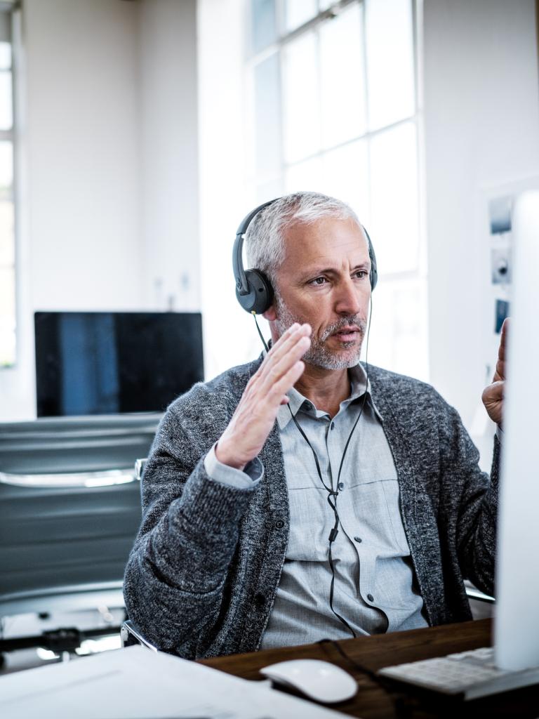 Een man met grijs haar zit te werken achter zijn computer. Hij draagt een koptelefoon.