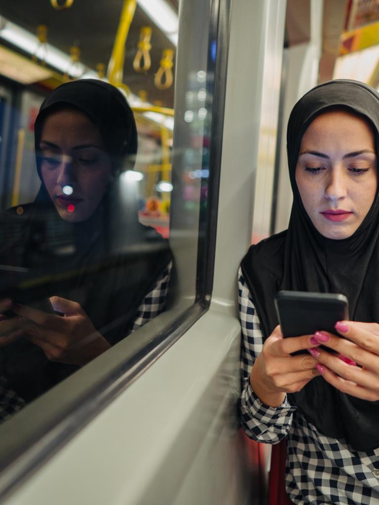Een vrouw van middelbare leeftijd met hoofddoek en geblokte blouse zit met haar telefoon in haar hand in het openbaar vervoer.