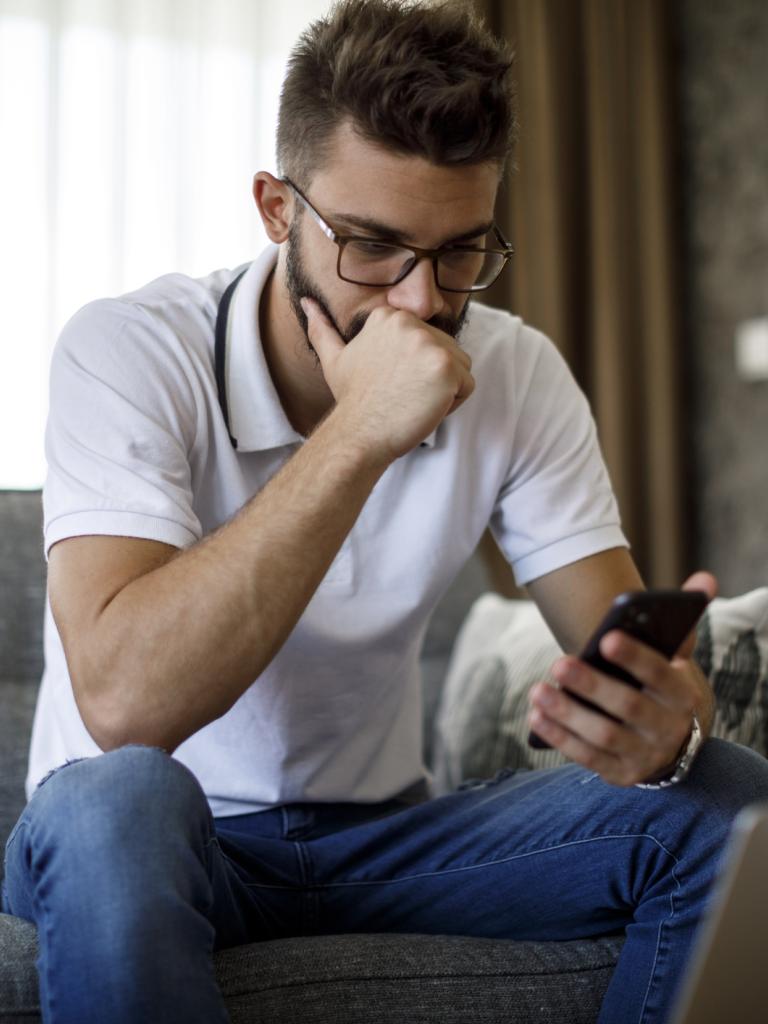 Jongeman met bril en wit t-shirt zit op zijn bank en kijkt ongerust naar zijn telefoon