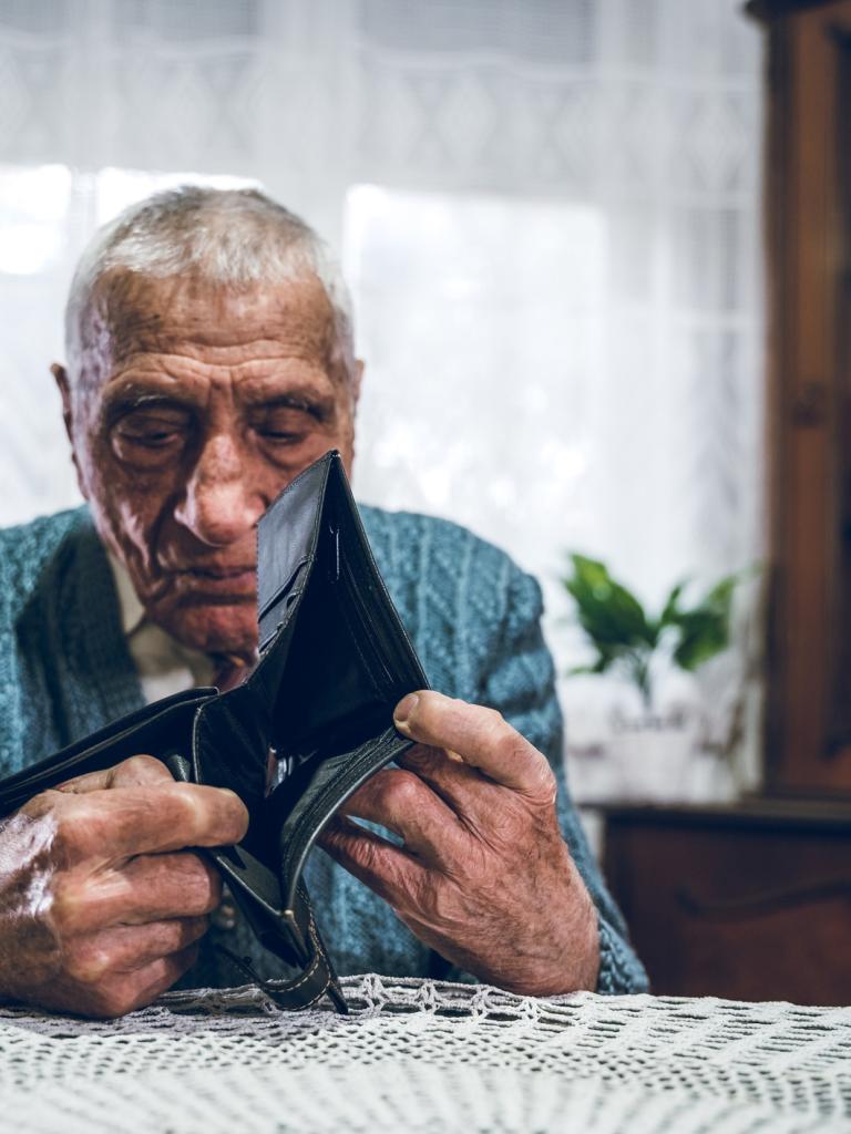 Een bejaarde man zit aan tafel en heeft een portemonnee in zijn handen. Er zit geen geld in de portemonnee.