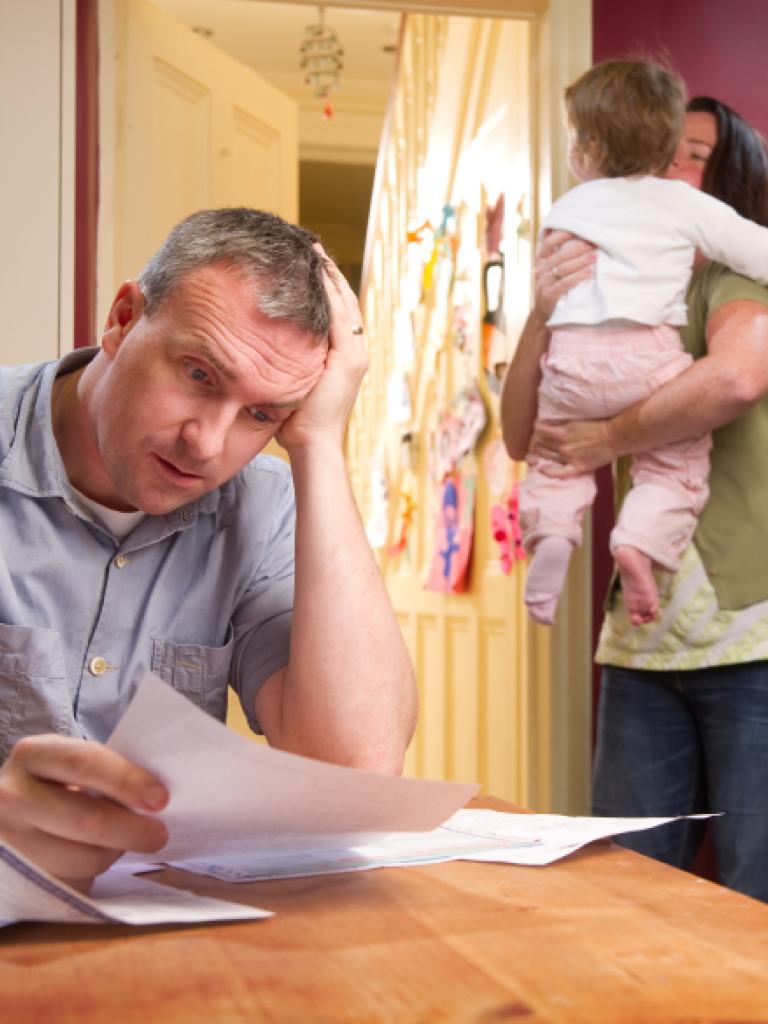 Foto van een bezorgde man aan een keukentafel en op de achtergrond zijn vrouw met hun kind in haar armen