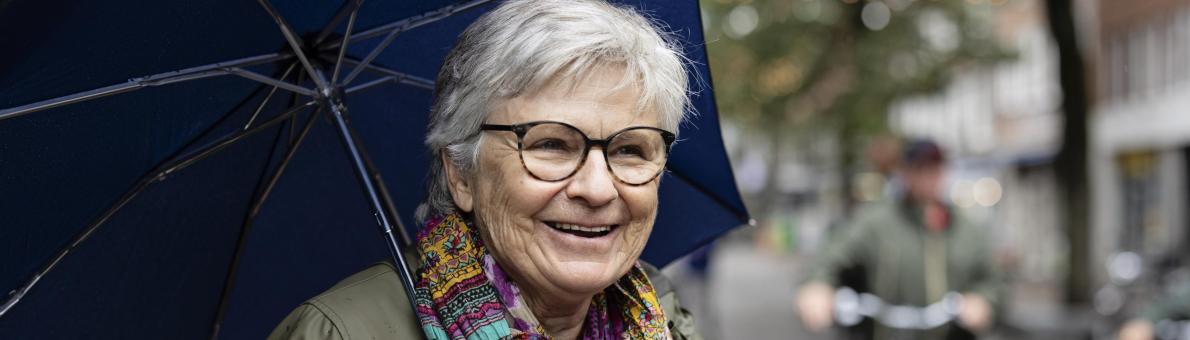 Marga, een oudere vrouw met grijs haar, bril en een gekleurde sjaal om. Ze heeft een paraplu vast Marga, een oudere vrouw met grijs haar, bril en een gekleurde sjaal om. Ze heeft een paraplu vast