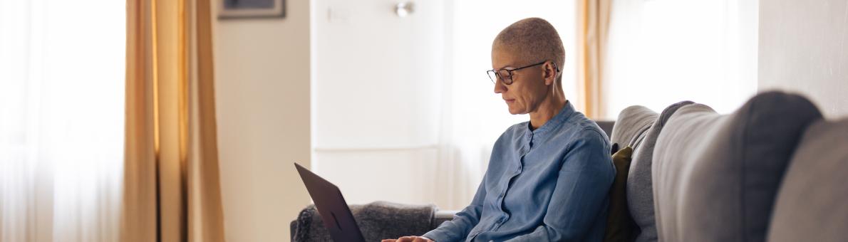 Een vrouw zonder haar zit met haar laptop op schoot in haar woonkamer op de bank.