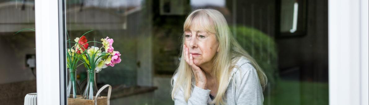 Oudere vrouw zit achter het raam en kijkt naar buiten. Ze kijkt zorgelijk en steunt haar hoofd op haar hand.