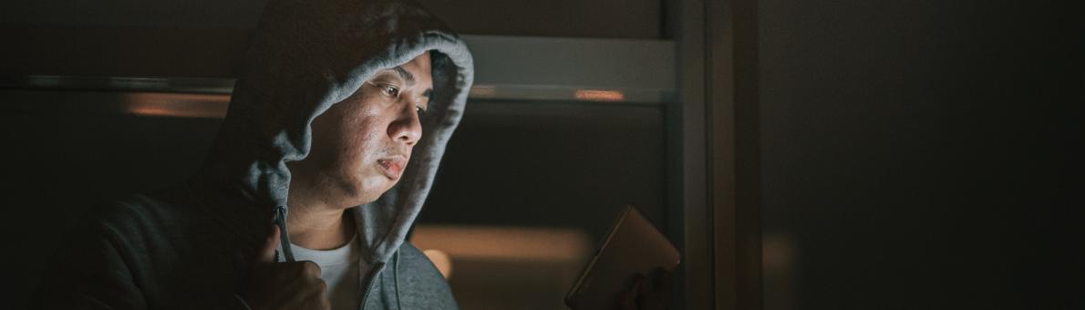 Een man zit in een donkere kamer, heeft zijn capuchon van zijn hoodie op en kijkt naar een tablet. Zijn gezicht is zichtbaar door het licht van de tablet.