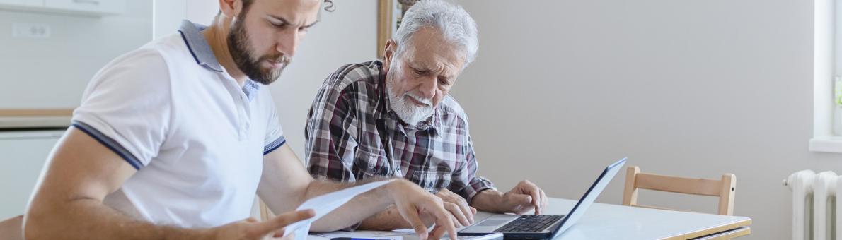 Een jongvolwassen man en senior man kijken samen naar de financiën. Ze hebben papierwerk, een rekenmachine en een laptop voor zich op tafel.