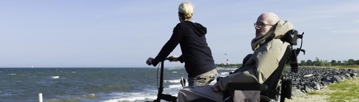 Een man in een rolstoel staat samen met een vrouw op de fiets bij het strand. Ze kijken naar de zee