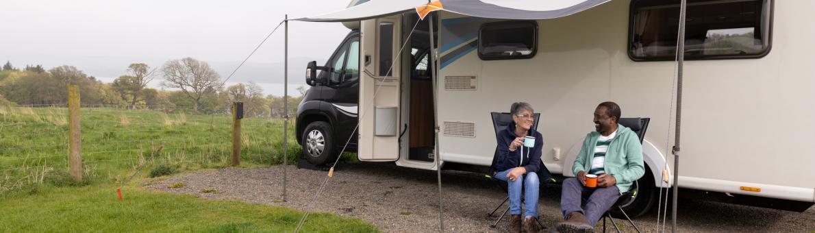 Een vrouw en een man zitten voor een camper op twee campingstoelen. Ze zijn in gesprek.