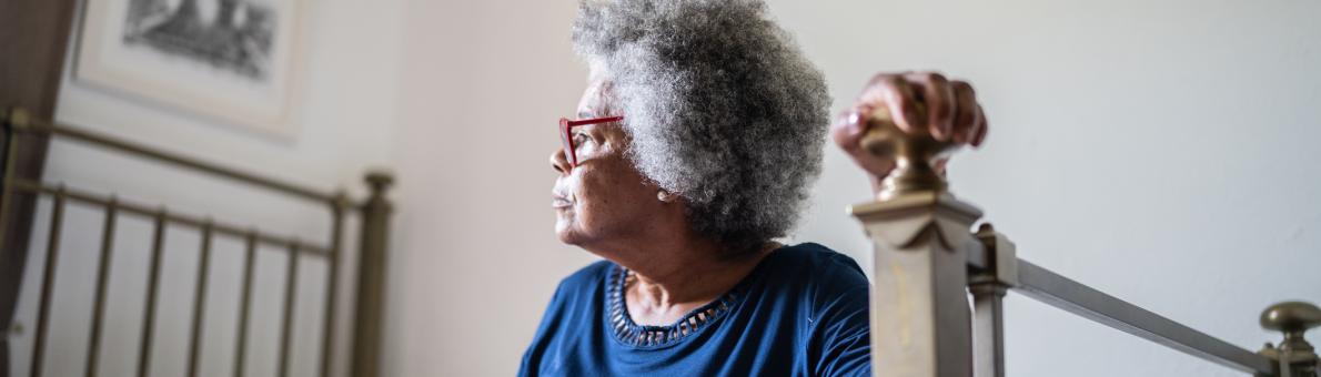 Senior vrouw met bril en blauw t-shirt zit op haar bed. Ze houdt met één hand het bedframe vast om op te gaan staan.