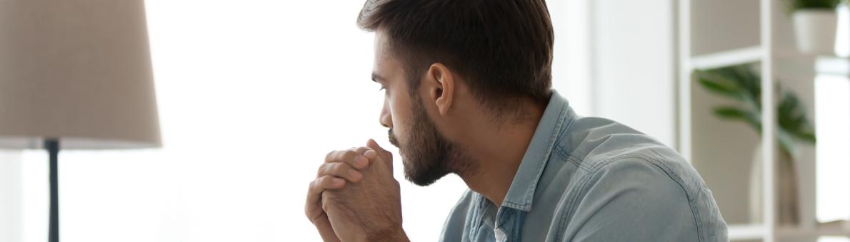 Een man van middelbare leeftijd met donker haar en een blauwe blouse zit op de bank. Hij kijkt nadenkend voor zich uit.