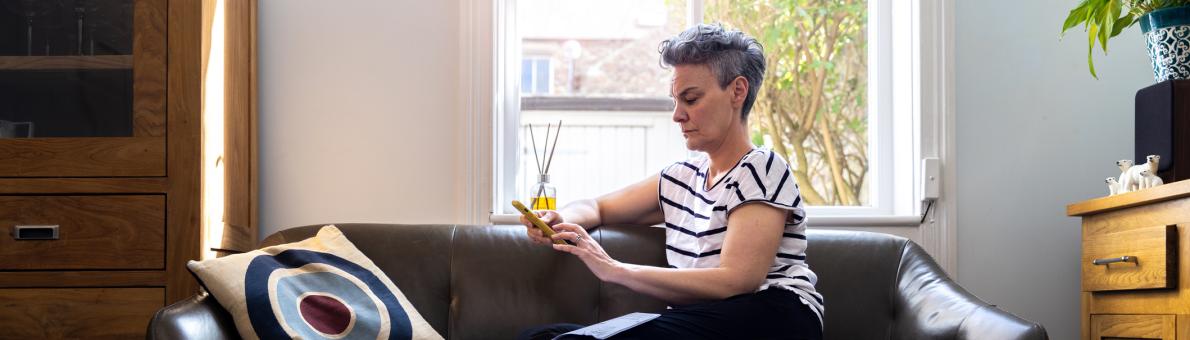 Vrouw zit thuis op haar bank voor het raam. Ze heeft haar telefoon vast en kijkt bezorgd.