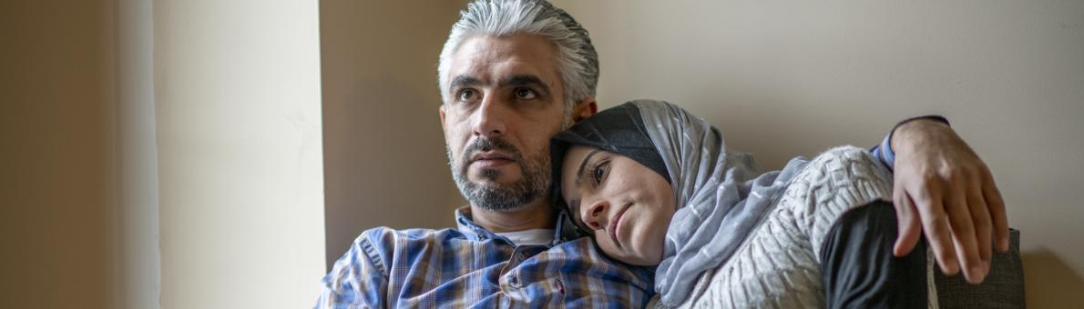 Een man en vrouw van islamitische afkomst zitten samen op de bank. Ze kijken bezorgd voor zich uit.