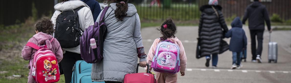 Groep mannen, vrouwen en kinderen lopen bij opvanglocatie. Ze dragen warme jassen, tassen en een rugzak. 