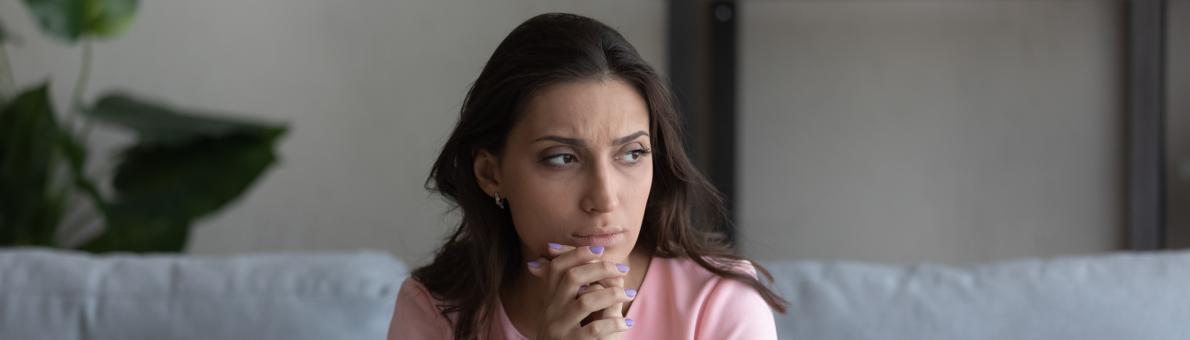 Vrouw met lang donker haar zit op de bank. Ze kijkt zorgelijk voor zich uit.