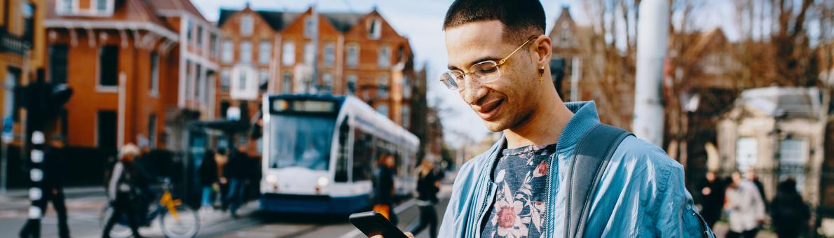 Een jongvolwassen man staat op straat met zijn telefoon in zijn hand. Op de achtergrond zie je mensen lopen en een tram.