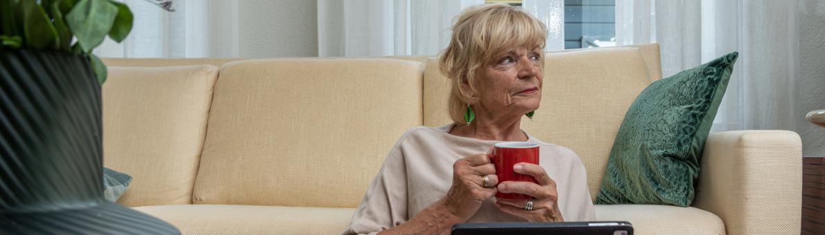 Oudere vrouw zit thuis op de grond voor haar bank. Ze kijkt opzij en houdt een kopje koffie vast.