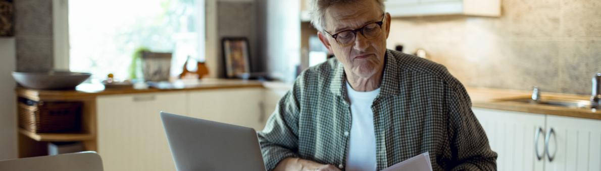 Senior man zit aan keukentafel met brief rekening in hand en laptop voor zich