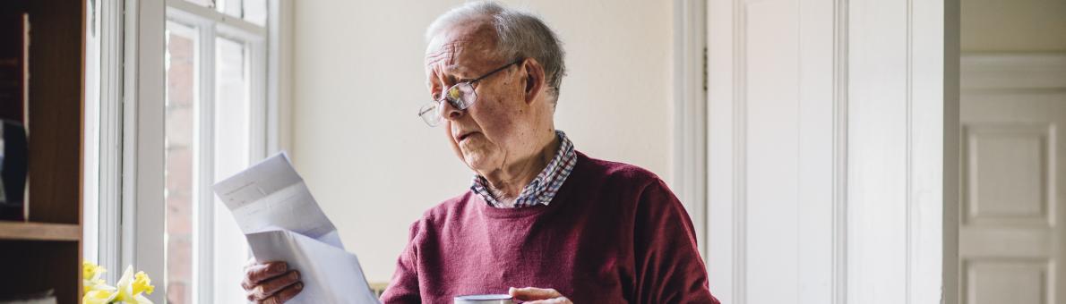 Senior man staat in de keuken met een brief in zijn ene hand en een kopje koffie in zijn andere hand.