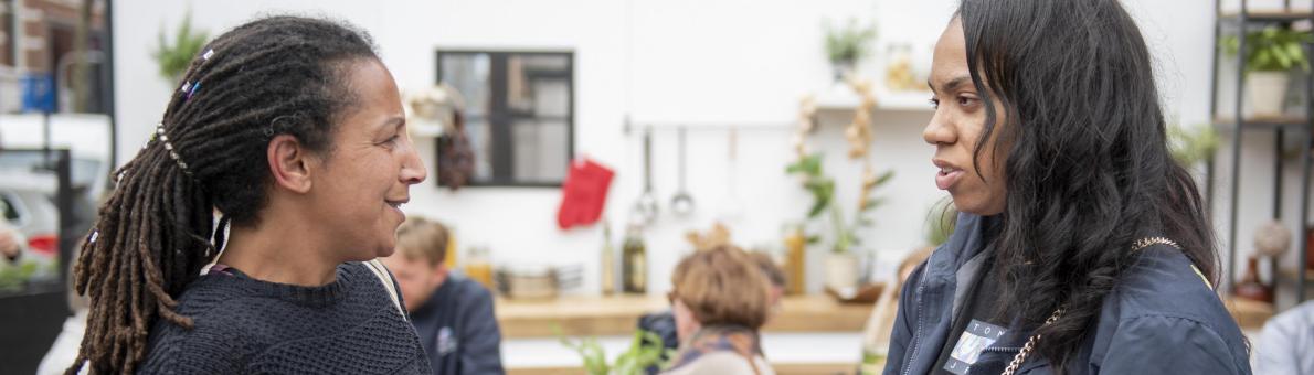 Twee vrouwen staan op een plein voor de pop-upkeuken van de Nationale ombudsman. Ze zijn in gesprek. 