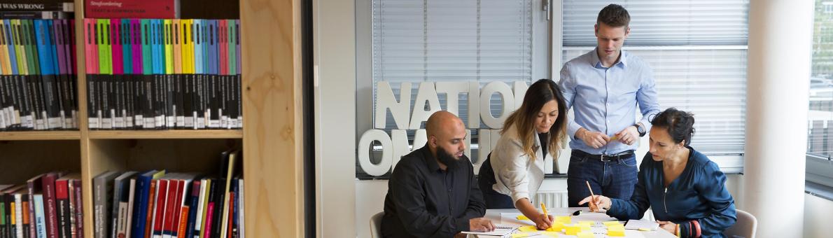 Medewerkers van de Nationale ombudsman zijn in gesprek. Ze zitten aan een tafel. Op de voorgrond staat een boekenkast Medewerkers van de Nationale ombudsman zijn in gesprek. Ze zitten aan een tafel. Op de voorgrond staat een boekenkast