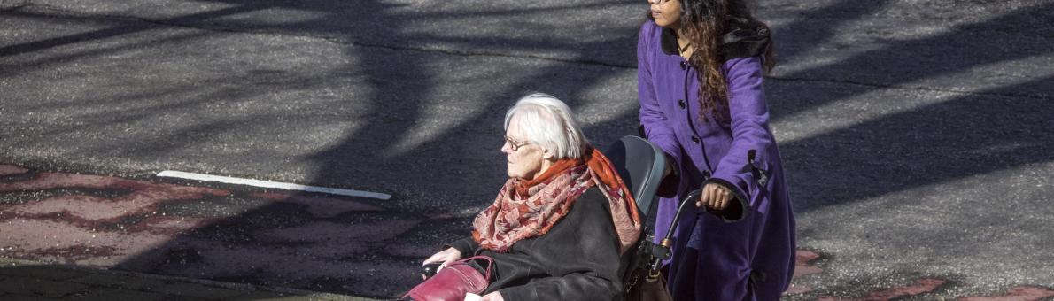 Mantelzorger (vrouw, lang donker haar en paarse jas) duwt rolstoel oudere vrouw (grijs haar, bril, donkere jas en rode sjaal