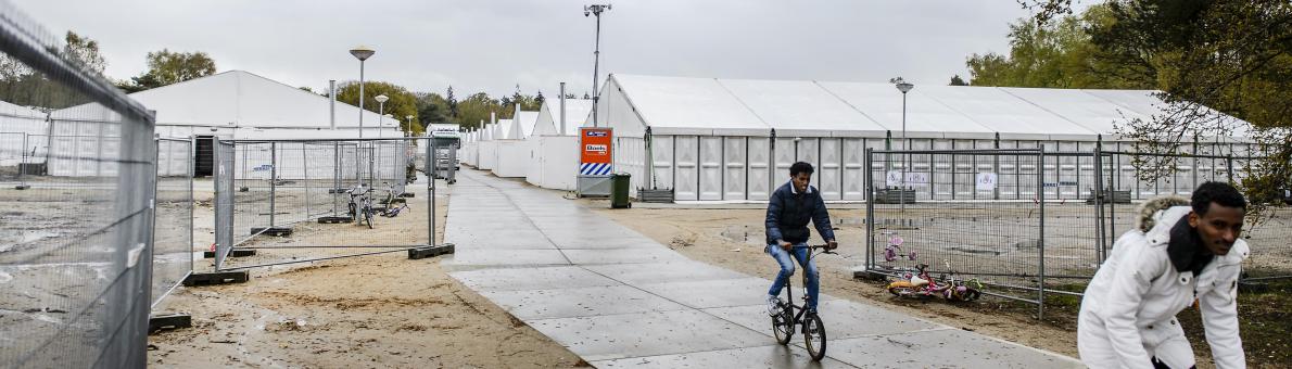 Twee mannen fietsen over het AZC terrein van Heumensoord. Tenten op de achtergrond